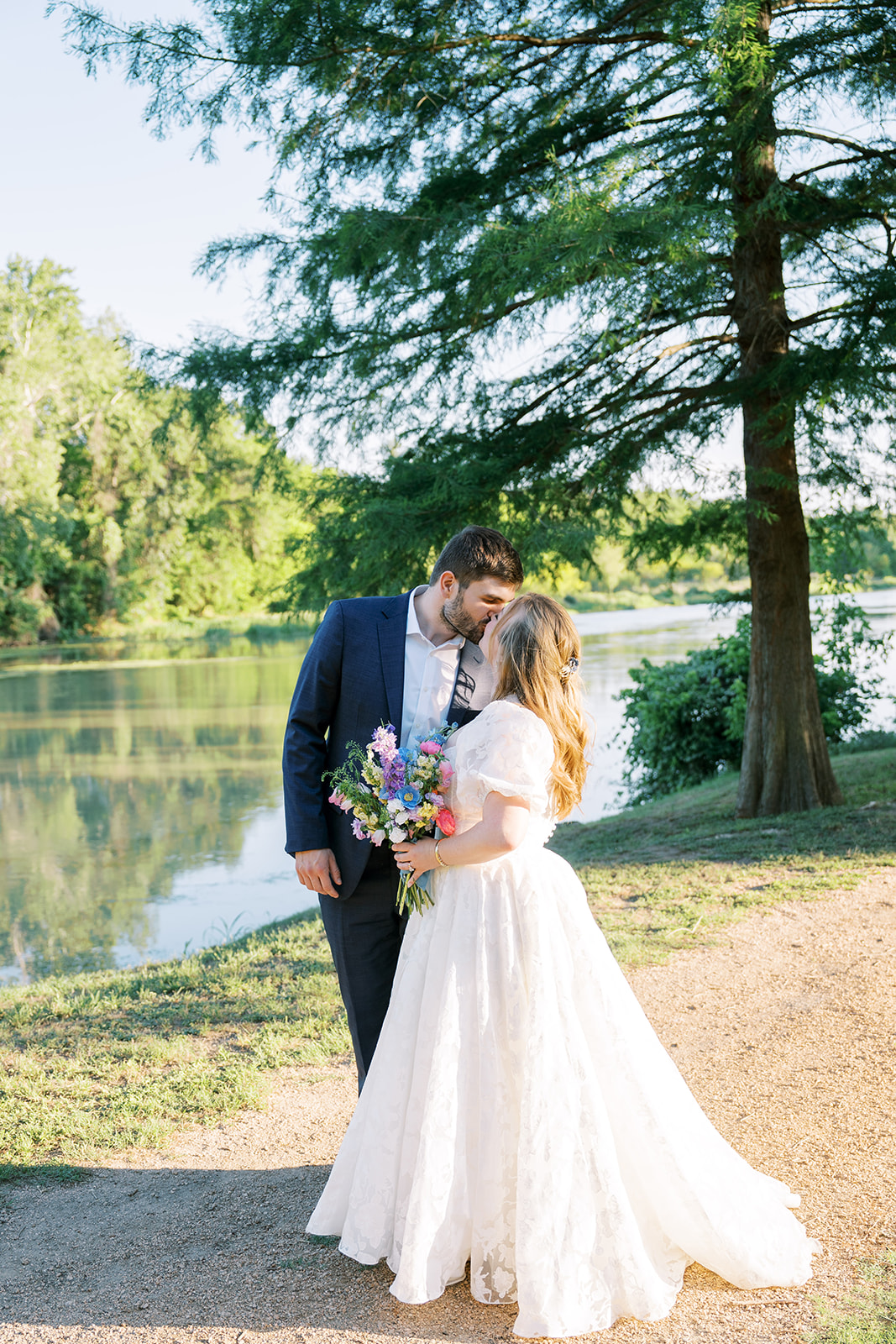 Bride and groom kiss in front of lake with lots of trees. Bride holds an Austin wedding flower bouquet