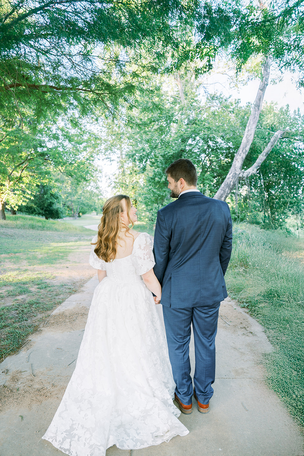 Bride and groom walk away from the camera holding hands looking at each other.
