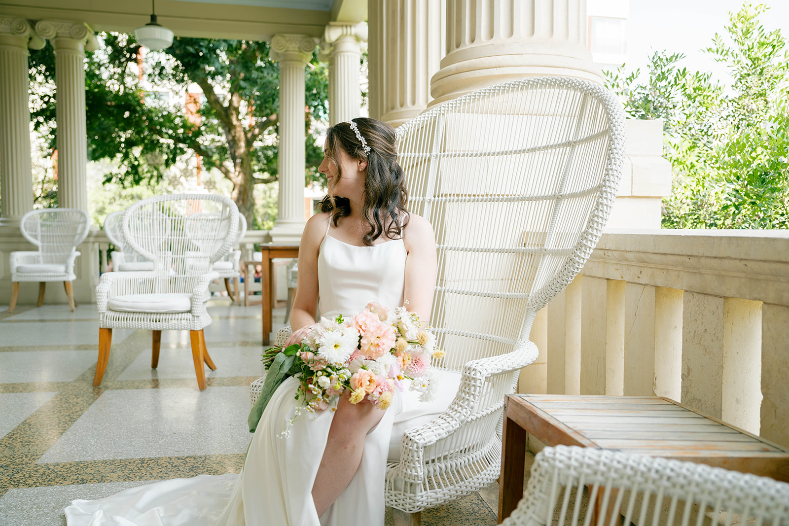 Bride sits on chair on the porch of Hotel Ella holding a bouquet of flowers looking away from the camera