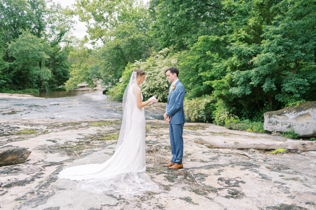 Bride and groom stand next to a river reading private vows with green trees behind them