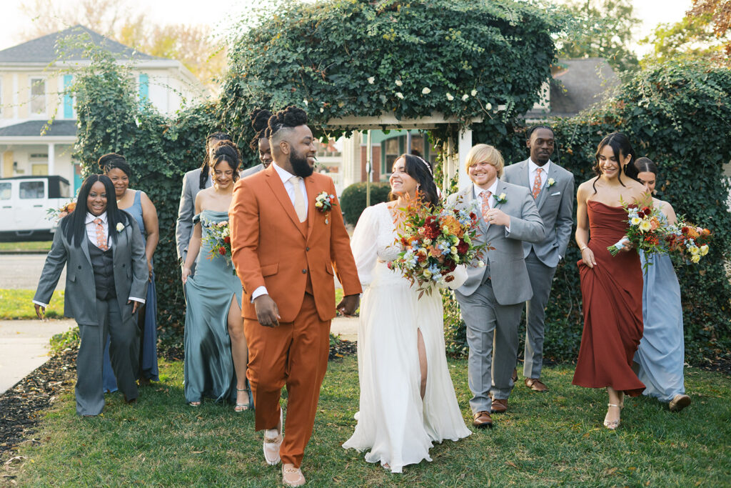 Bride and groom hold hands walking in front of bridal party with a beautiful arch and greenery. Time for bridal party photos on wedding day timeline