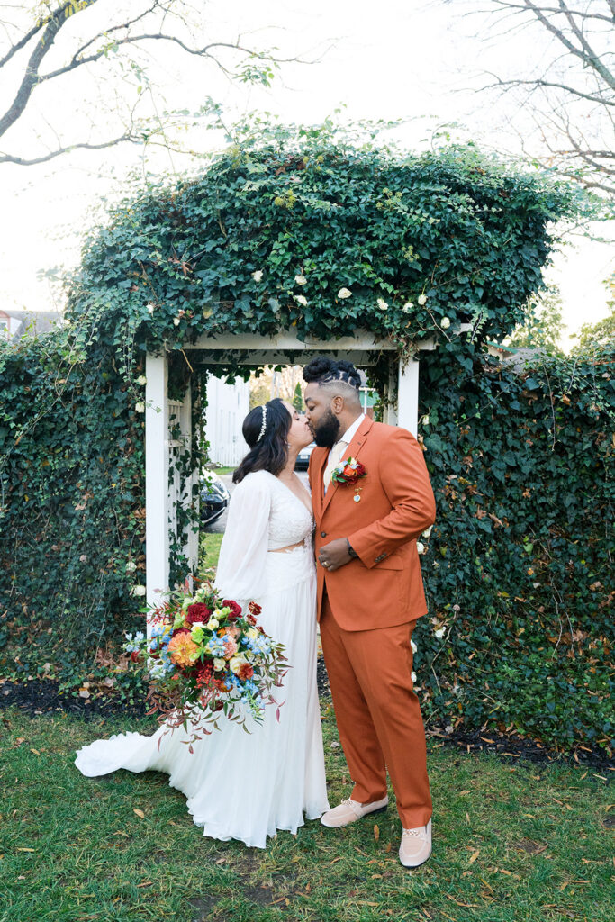 Bride holds red, orange, blue, white flower bouquet kissing groom wearing orange suit. They stand in front of a wedding arch covered in greenery. Couple photos added to wedding day timeline