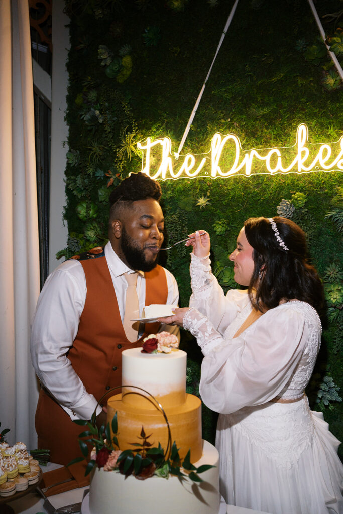 Bride feeds groom cake in front of their name sign the cake is white on the top and bottom layers and light orange in the middle layer. Cake cutting added to wedding day timeline